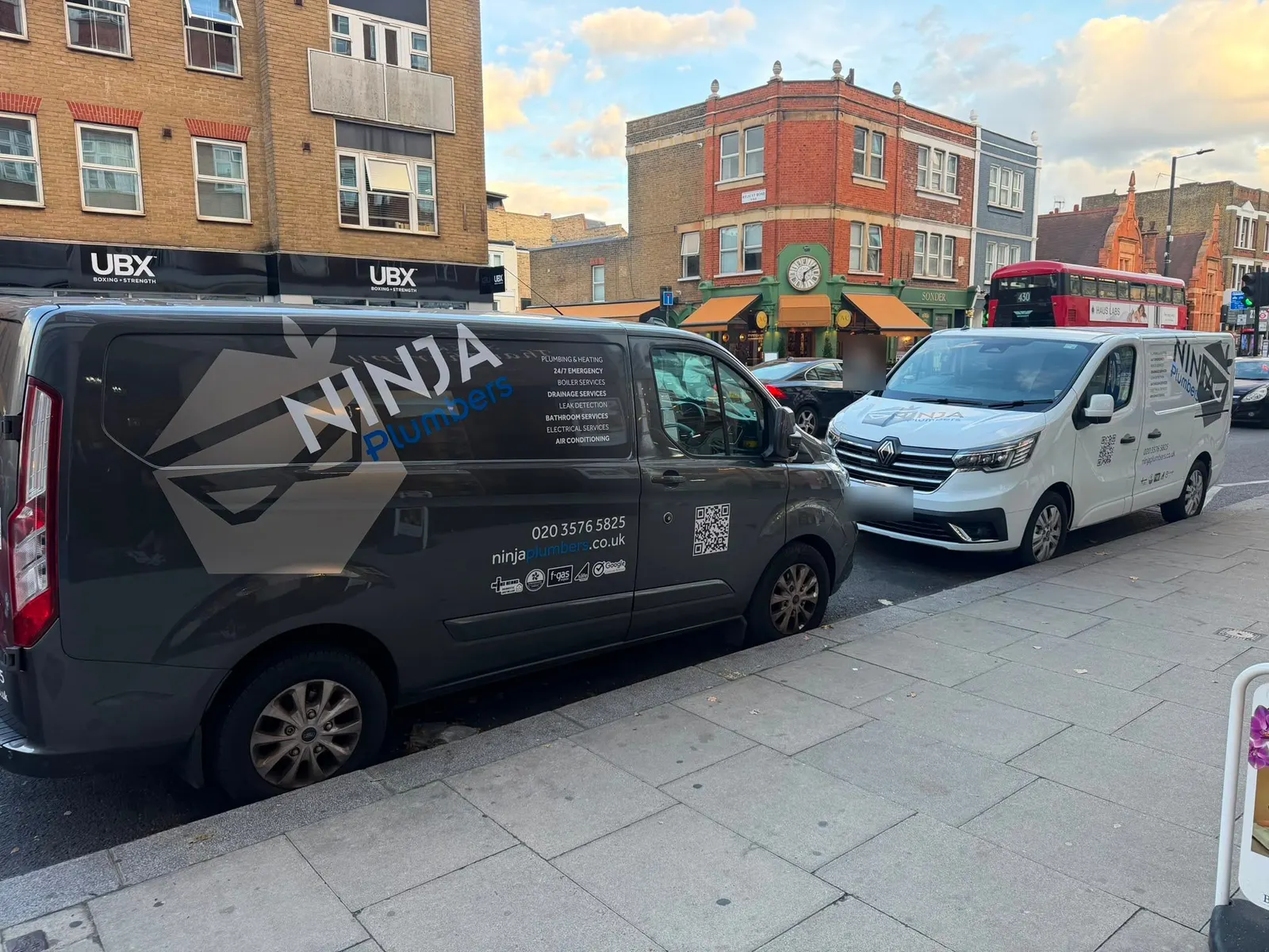 Two Ninja Plumbers vans, one dark grey and one white, parked on a London high street with the company phone number and website on the side